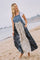 woman wearing a denim overall dress with a patterned skirt, standing on the beach near the water