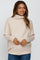 woman wearing a soft beige maternity sweater with a cowl neck and long sleeves, gently smiling and showing her baby bump
