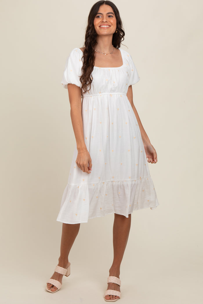 woman wearing a white puff sleeve midi dress with floral embroidery and a tiered hem, smiling and standing against a neutral background