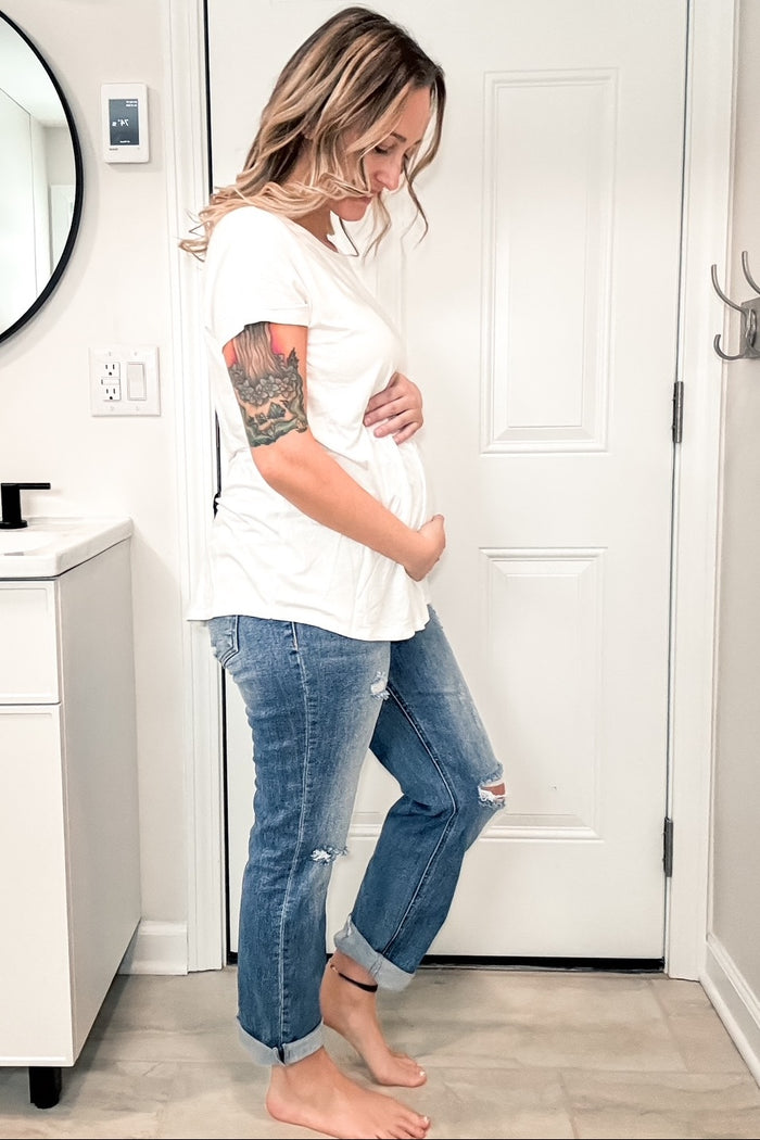 woman wearing a white short sleeve maternity top and distressed blue jeans, holding her baby bump