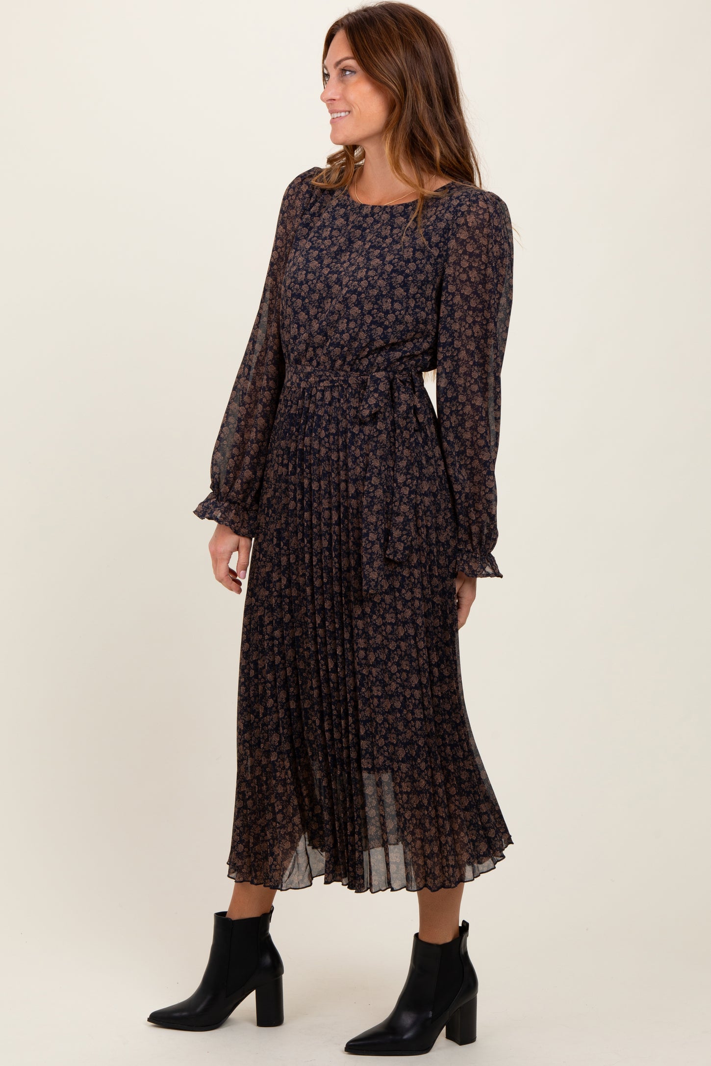 woman wearing a dark floral long sleeve midi dress with a belted waist and pleated skirt, paired with black ankle boots