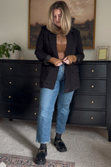 woman wearing a black blazer over a brown knit top and blue denim jeans, styled with black loafers and wavy hair