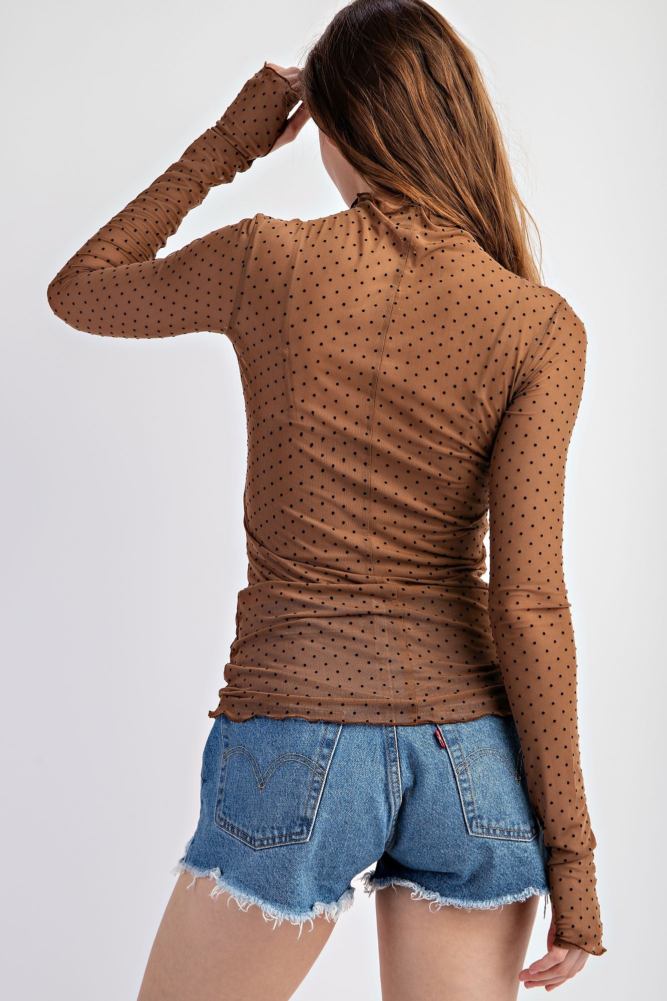 woman wearing a brown polka dot mesh long sleeve top paired with denim shorts, showing the back view