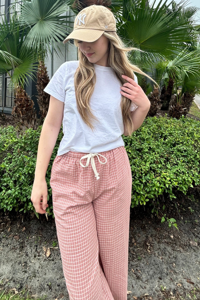 woman wearing a white t-shirt and pink checked wide-leg pants, standing outdoors with palms in the background