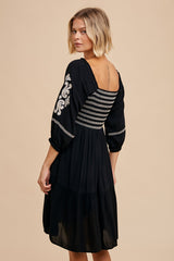 woman wearing a black embroidered dress with three-quarter sleeves and a tiered skirt, viewed from the back