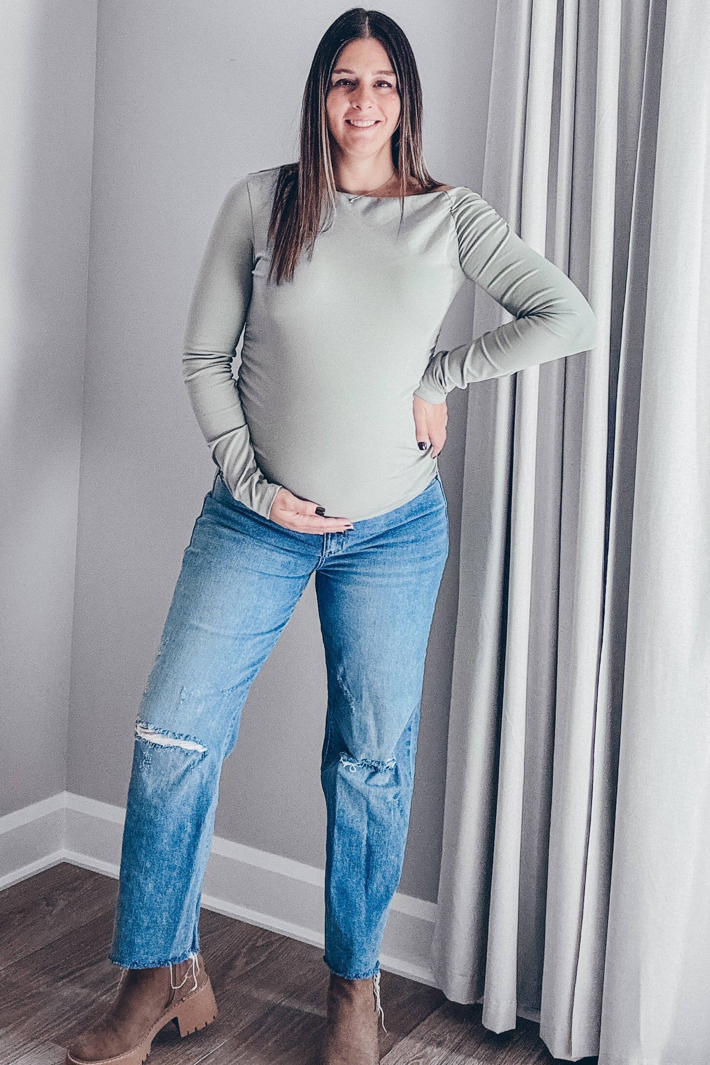 woman wearing a light green long sleeve maternity top and distressed jeans, holding her baby bump