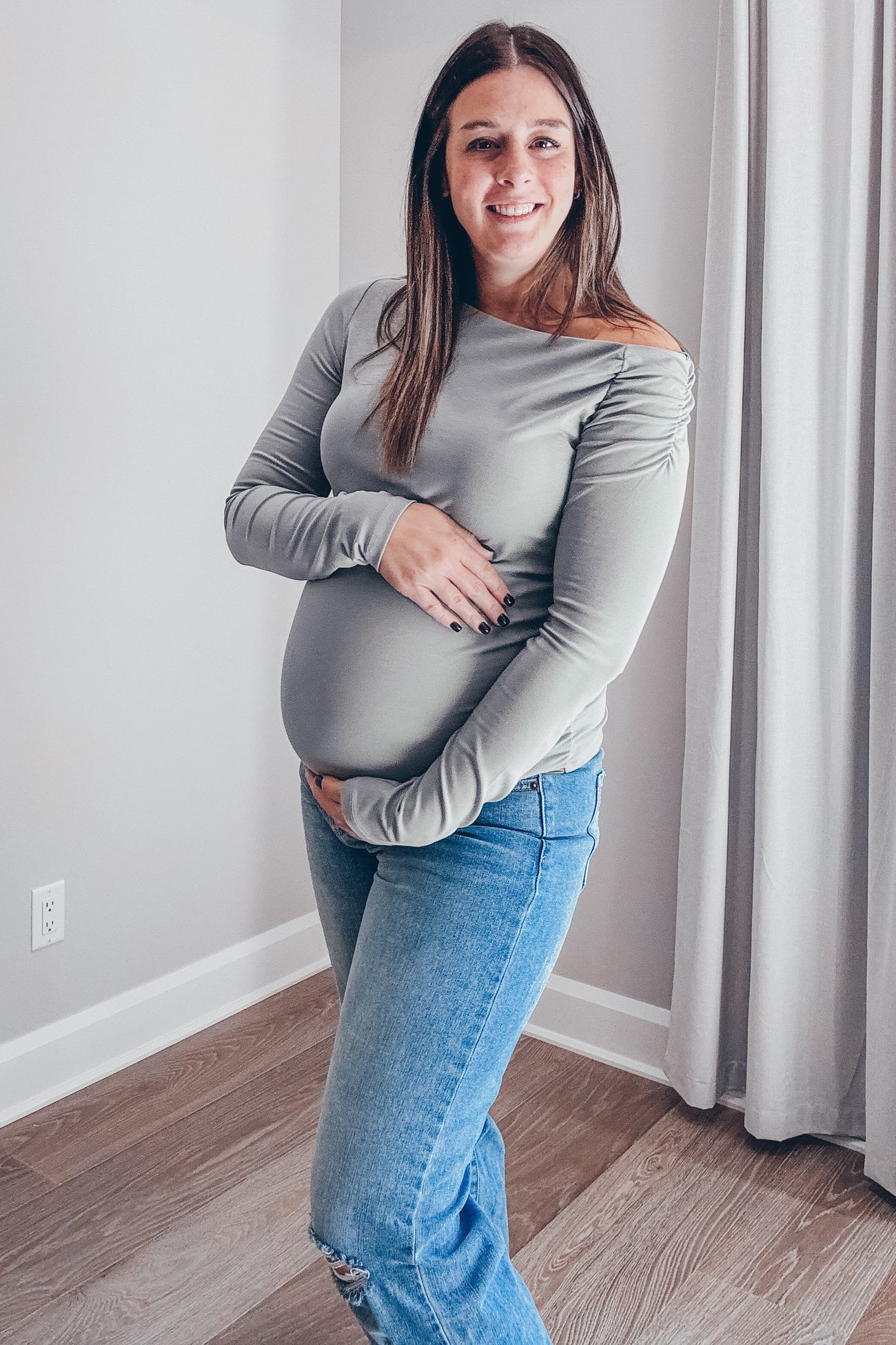 woman wearing a gray off-the-shoulder maternity top and holding her baby bump, paired with distressed blue jeans