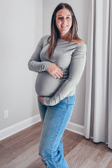 woman wearing a gray off-the-shoulder maternity top and holding her baby bump, paired with distressed blue jeans