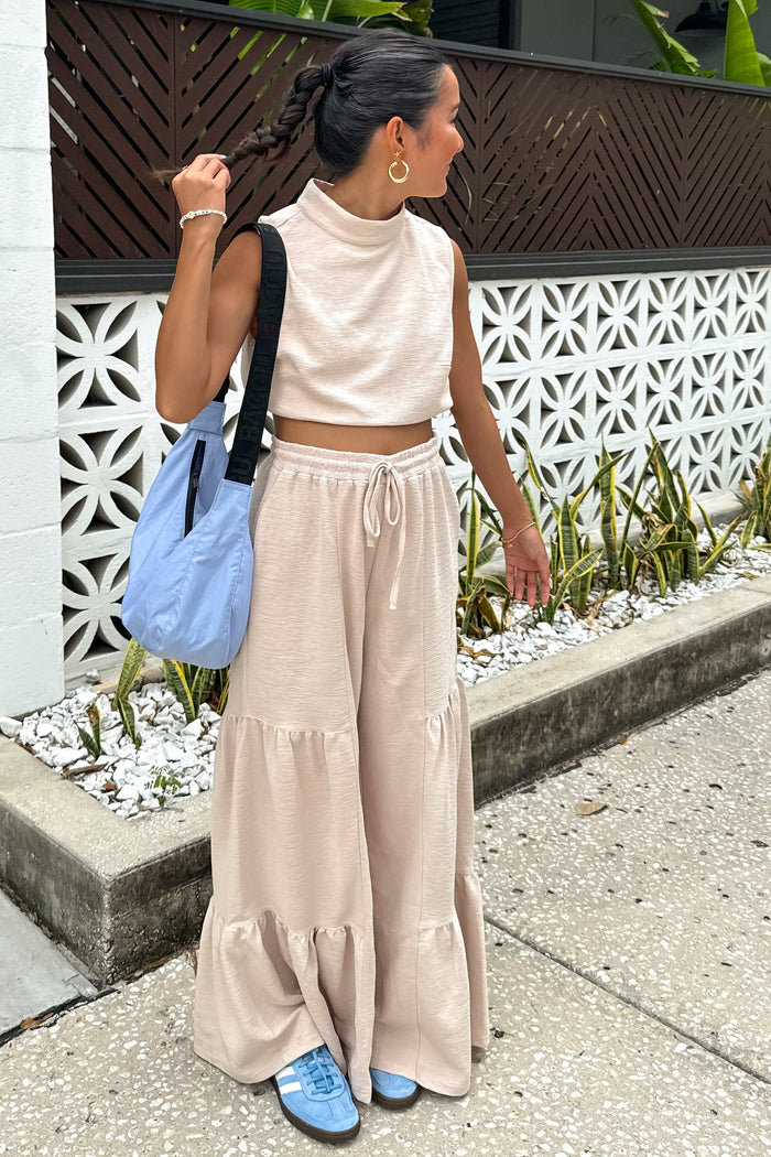 woman wearing a beige cropped sleeveless top and matching tiered wide-leg pants, carrying a light blue bag