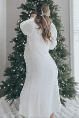 woman wearing a white maternity dress with long sleeves, standing with a baby bump in front of a decorated Christmas tree