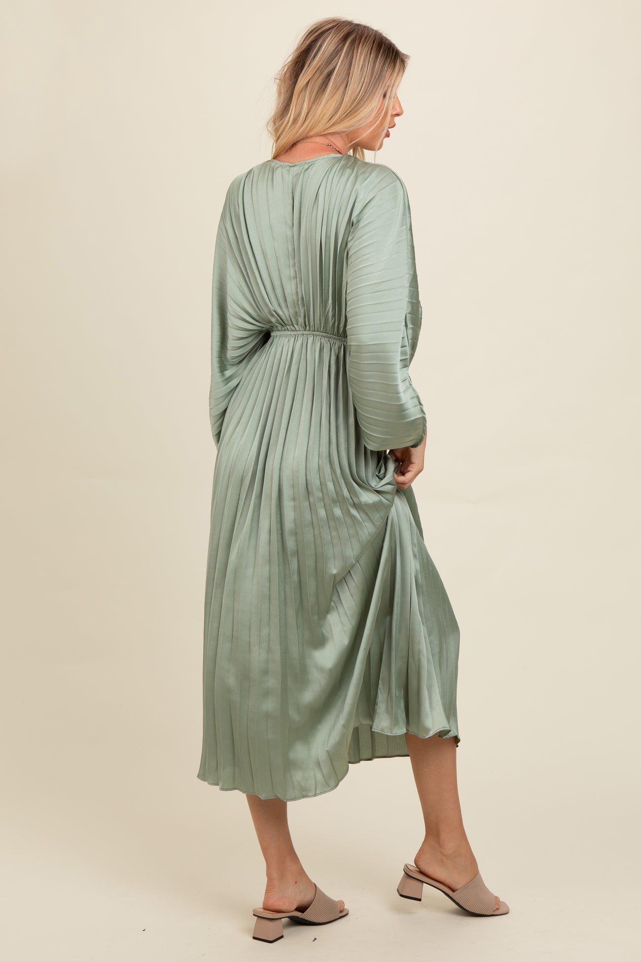 woman wearing a green pleated midi dress with long sleeves and a cinched waist, standing sideways
