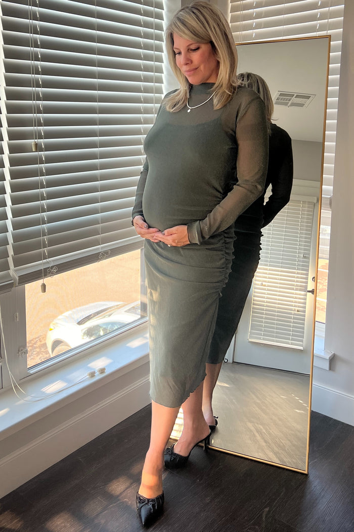 woman wearing a dark green long sleeve maternity dress, gently cradling her baby bump while standing by a mirror