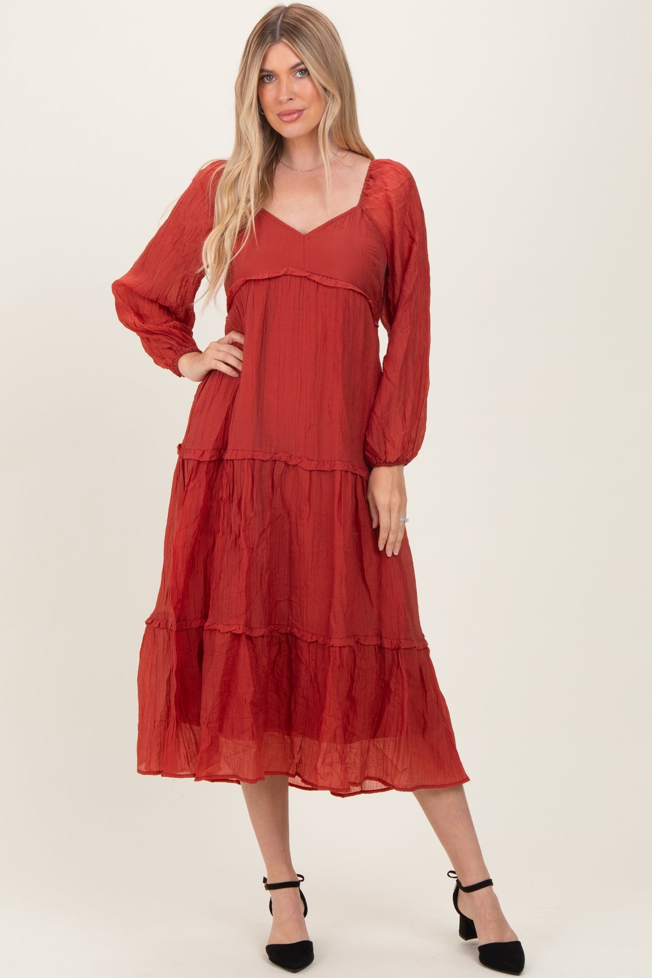 woman wearing a rust red long sleeve maxi dress with a tiered skirt and v-neckline, posing against a neutral background