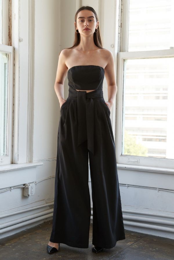woman wearing a black strapless jumpsuit with a tied waist and wide-leg pants, standing against a bright window