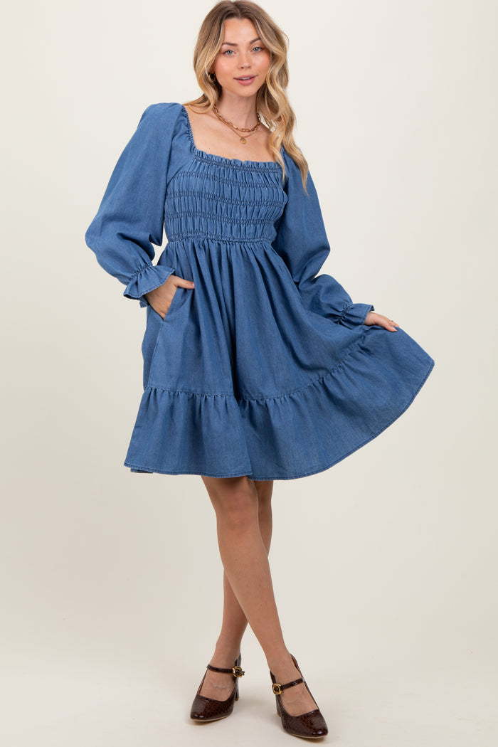 woman wearing a blue denim smock dress with long puff sleeves and ruffle hem, standing with one hand in pocket