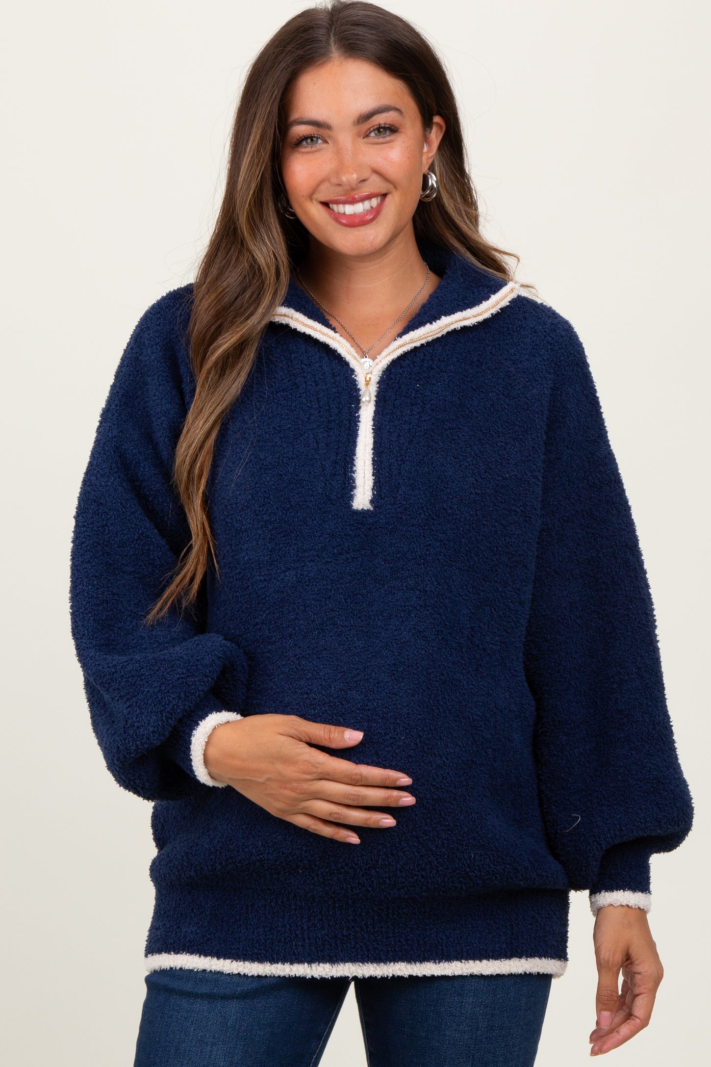 woman wearing a navy blue fuzzy maternity pullover with a quarter zip and white trim, holding her baby bump