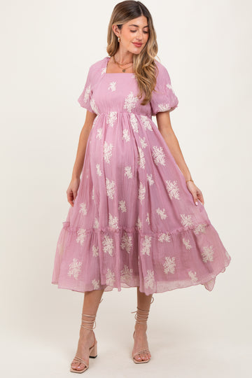 woman wearing a pink maternity dress with floral embroidery and a tiered skirt, showcasing her baby bump
