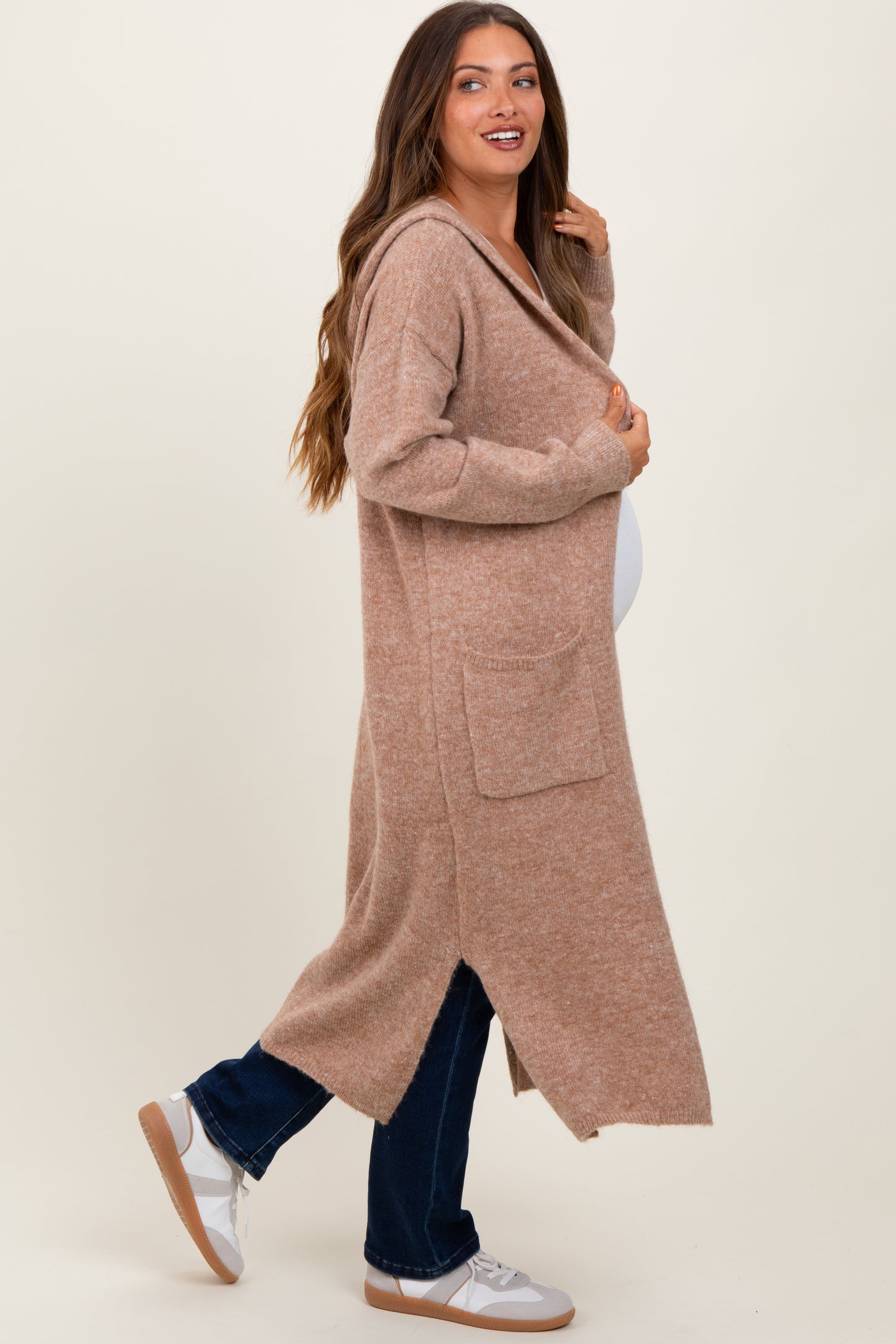 woman wearing a tan maternity cardigan with pockets and a side slit, holding her baby bump while smiling