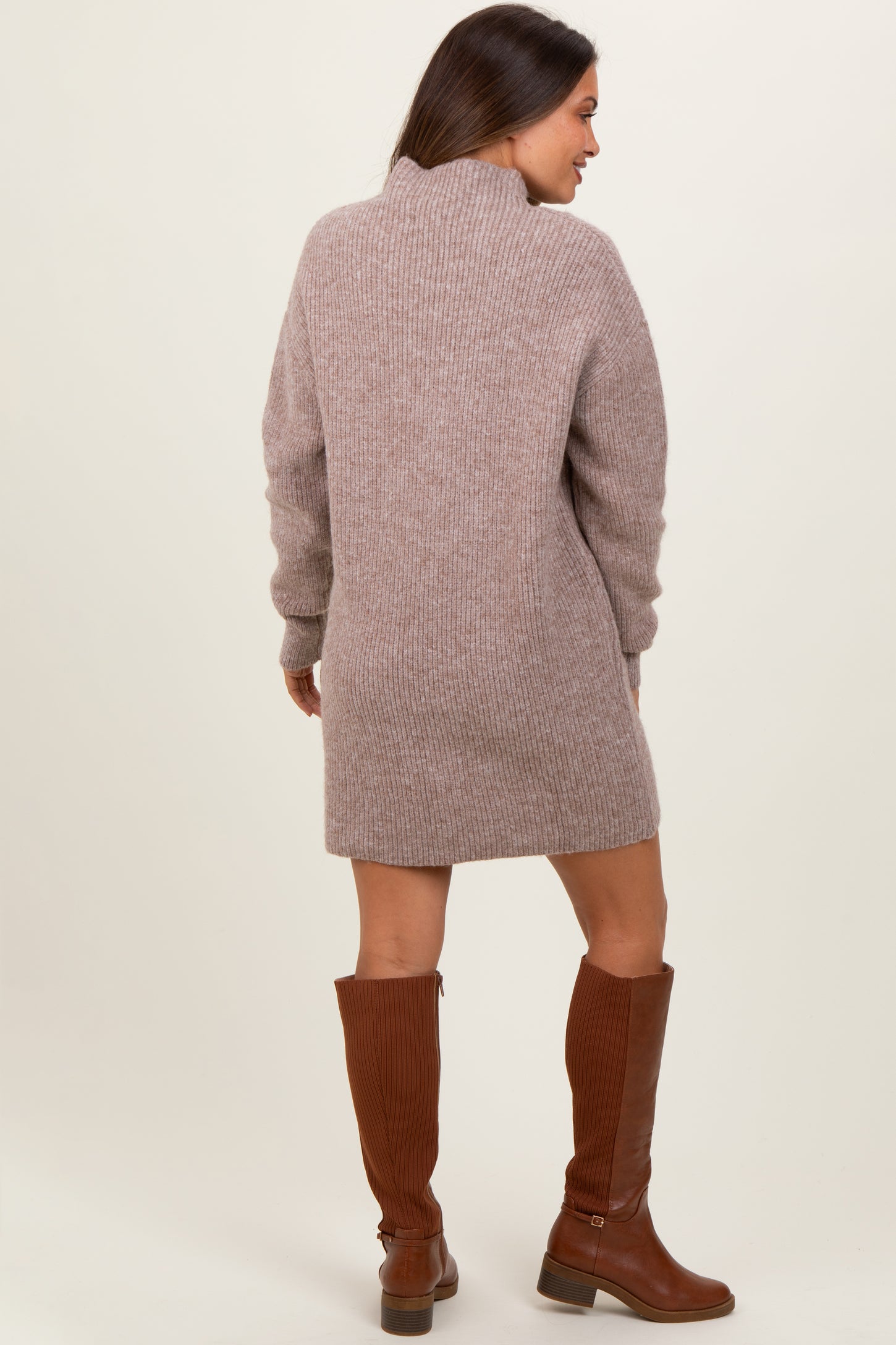 woman wearing a taupe knit maternity sweater dress with a high collar and long sleeves, paired with brown knee-high boots