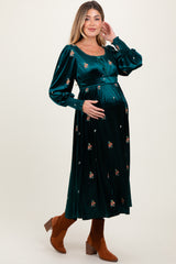 woman wearing a deep green velvet maternity dress with floral embroidery and long sleeves, visible baby bump