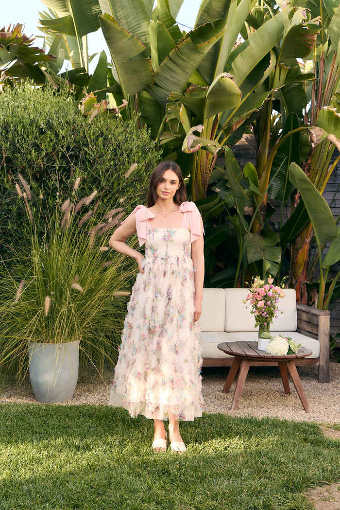 woman wearing a light pink floral maxi dress with ruffled layers and bow details on the shoulders in a garden setting