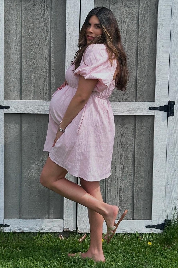 woman in a light pink short sleeve maternity dress showing her baby bump while standing next to a wooden fence