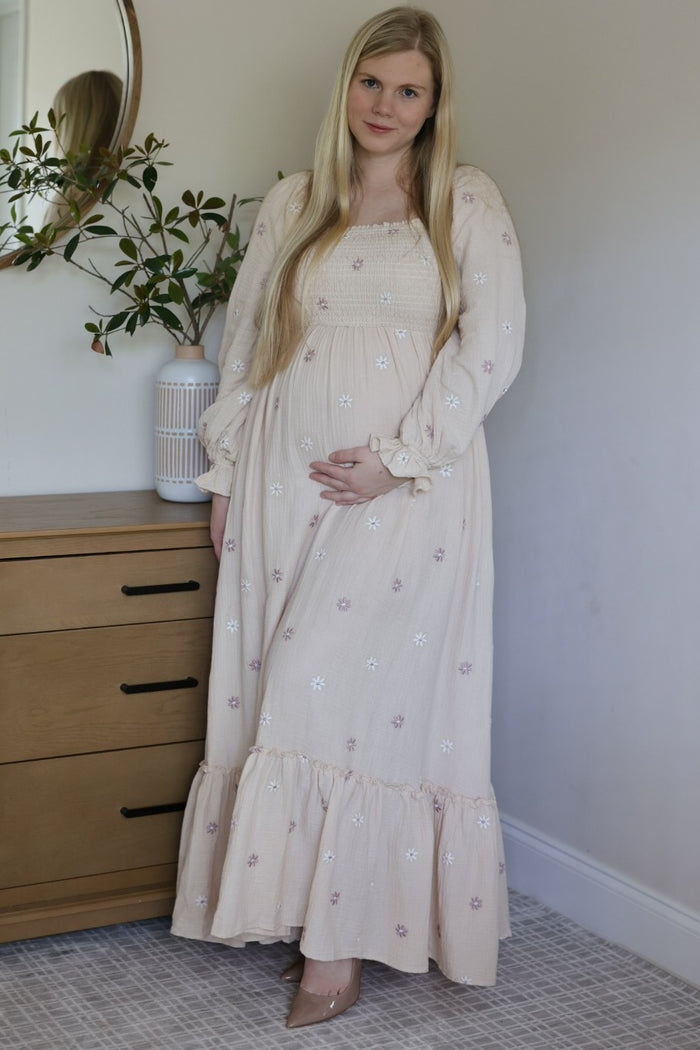 woman wearing a light pink maternity dress with floral embroidery and smocked bodice, holding her baby bump