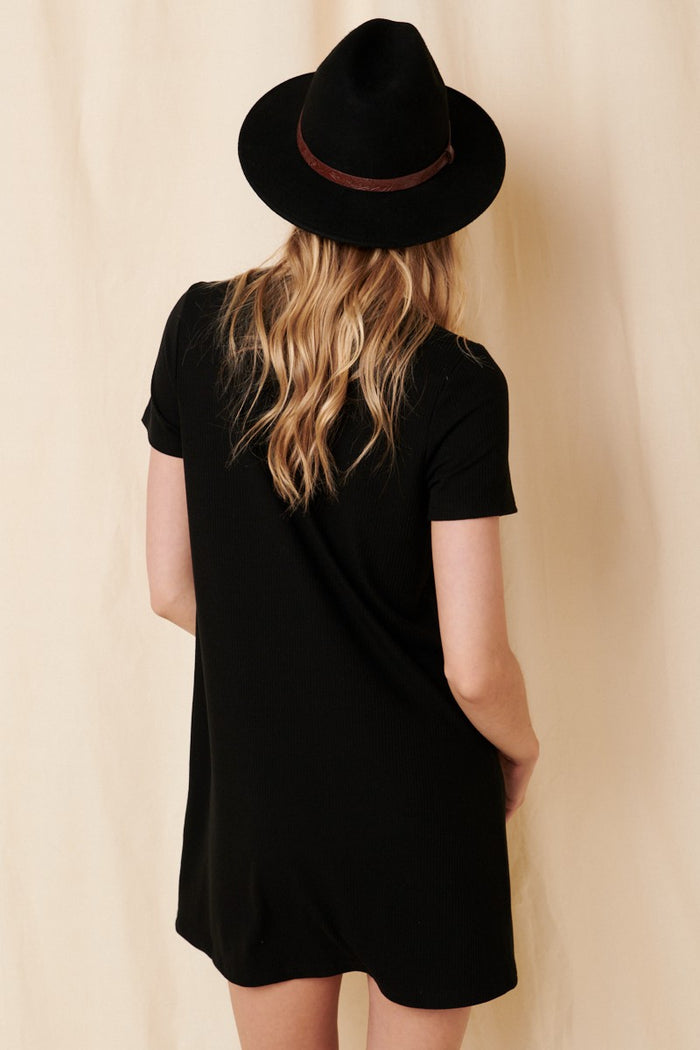woman wearing a black short sleeve dress and a black hat, viewed from the back against a neutral background