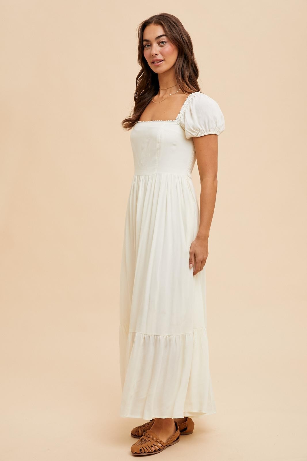 woman wearing a cream-colored maxi dress with puff sleeves and a square neckline, posing against a light background