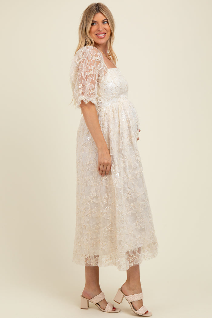 woman wearing a cream maternity dress with floral lace details and a baby bump, standing in profile