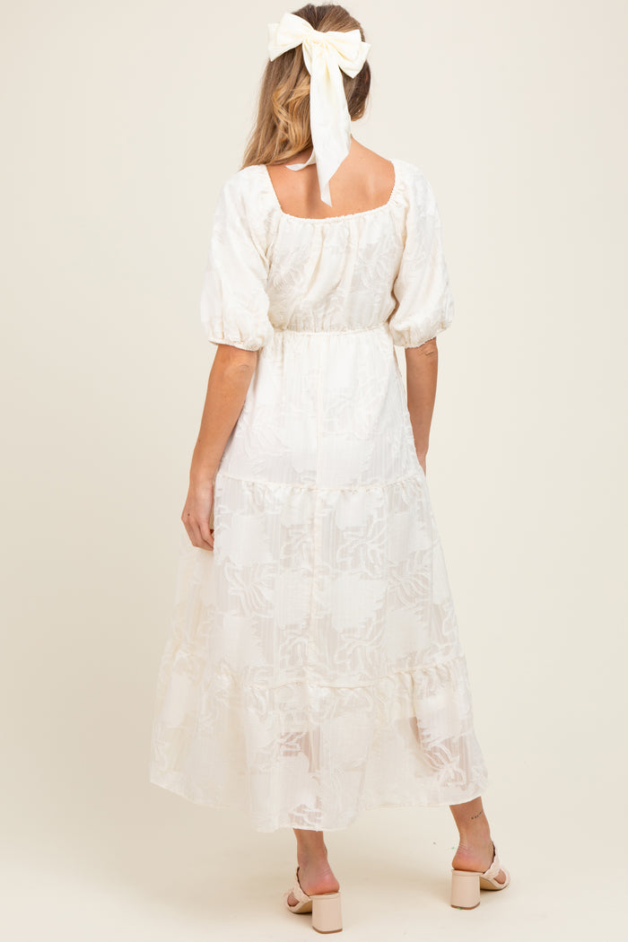 woman wearing a cream maternity maxi dress with puff sleeves and a tiered skirt, styled with a hair ribbon