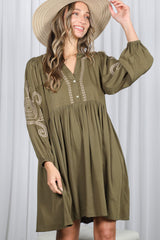 woman wearing a green long sleeve dress with embroidered details and a buttoned front, holding a wide-brimmed hat