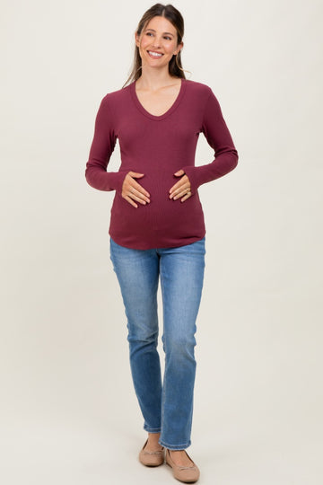 woman wearing a burgundy long sleeve maternity top and holding her baby bump, paired with blue jeans