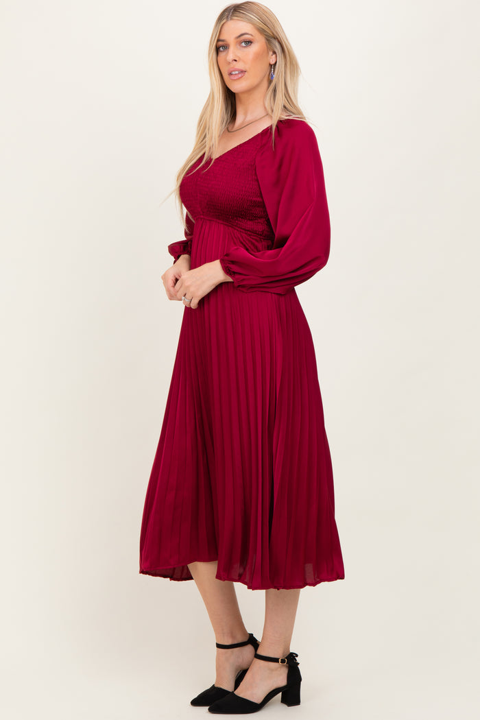 woman wearing a burgundy pleated midi dress with long sleeves and a smocked bodice, standing sideways in profile