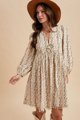 woman wearing a cream floral long sleeve dress with a tie detail and pom-pom trim, accessorized with a brown hat