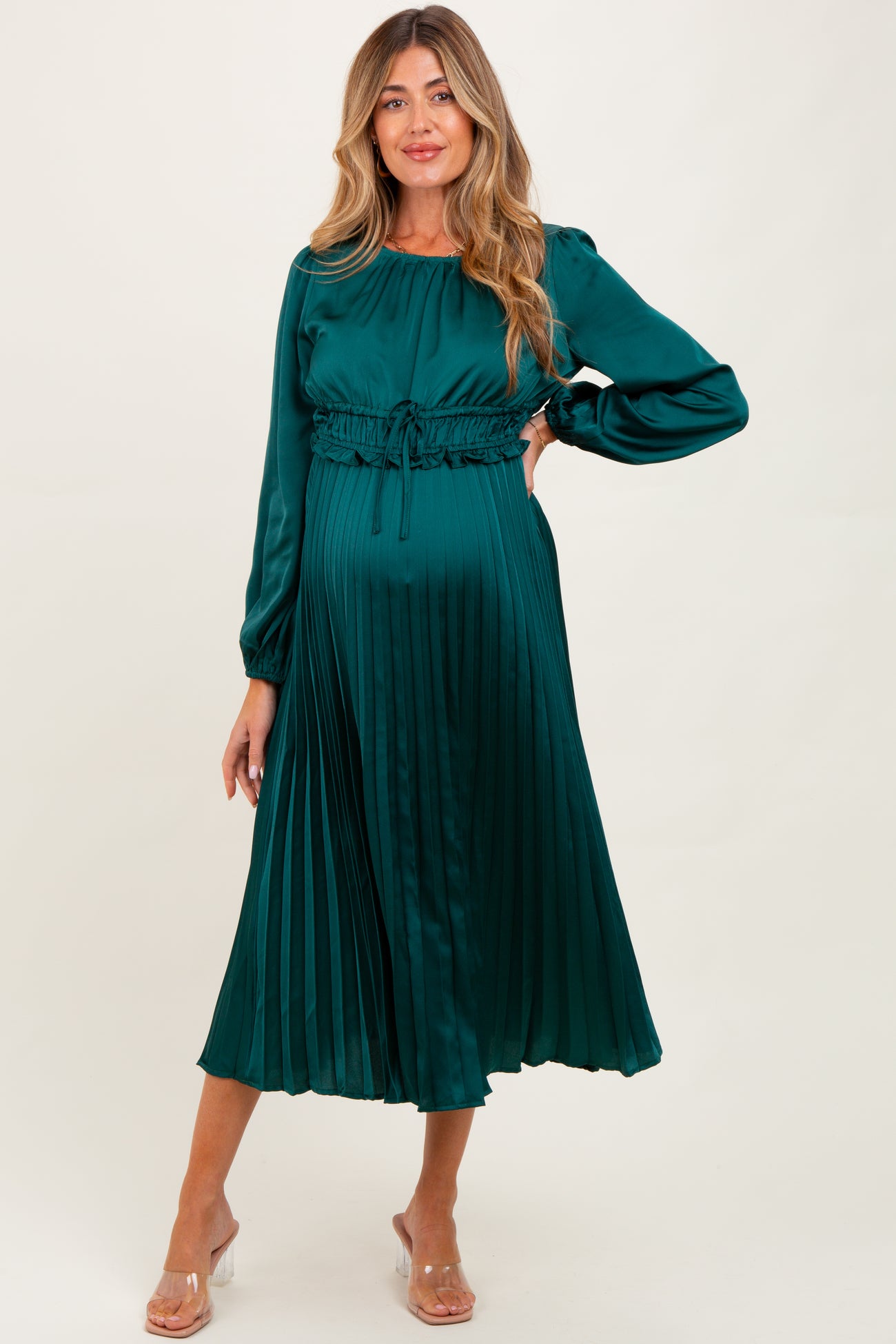 woman wearing a teal pleated maternity dress with a cinched waist and long sleeves, holding her baby bump