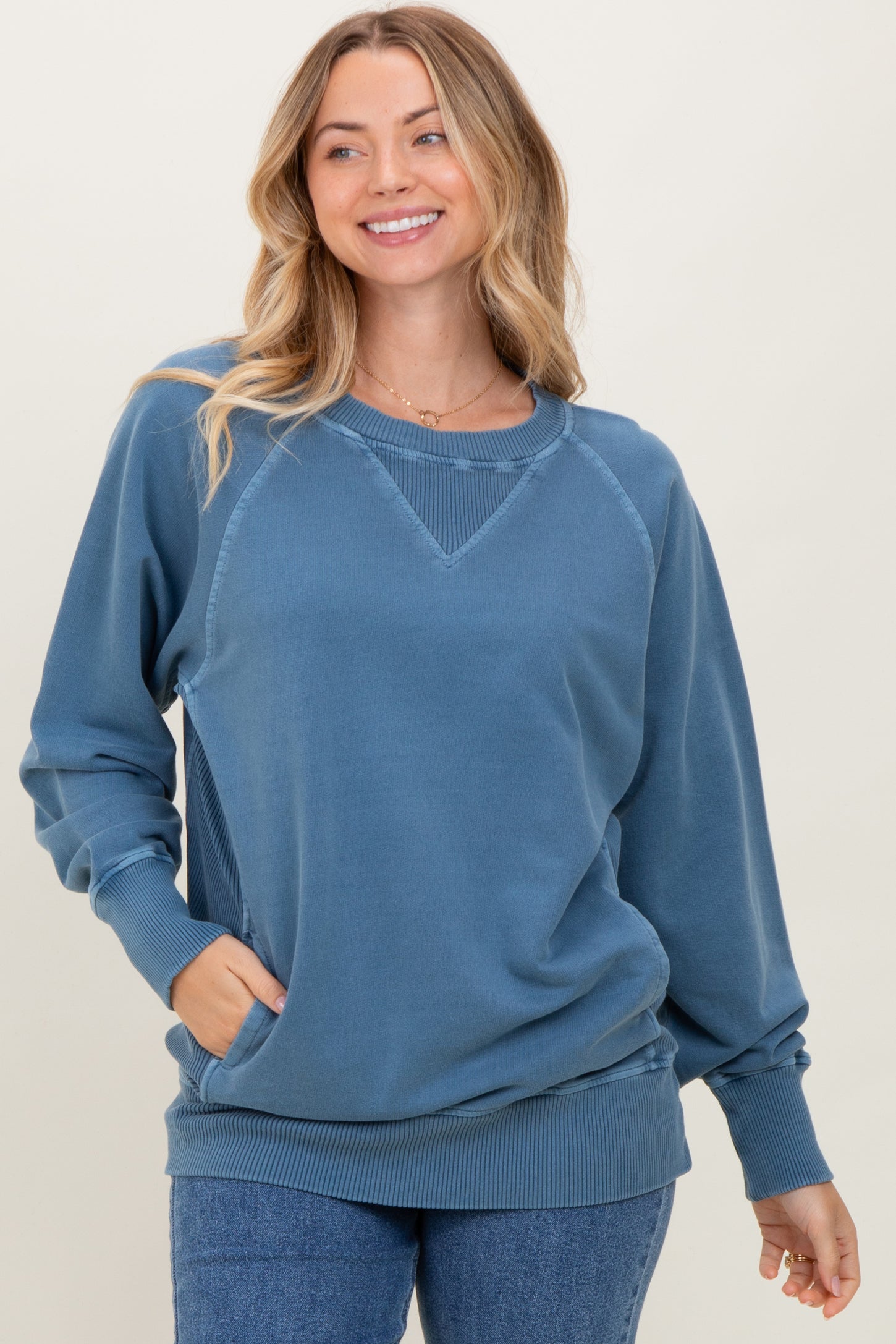 woman wearing a blue oversized sweatshirt with a round neckline and ribbed cuffs, smiling against a neutral background