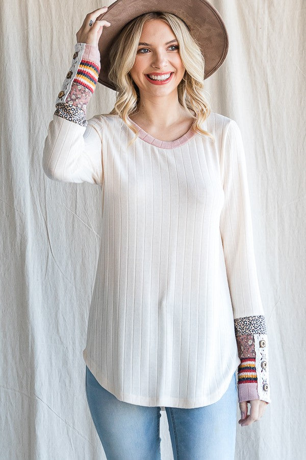 woman wearing a cream long sleeve top with patterned sleeves and a round neckline, smiling while holding a hat