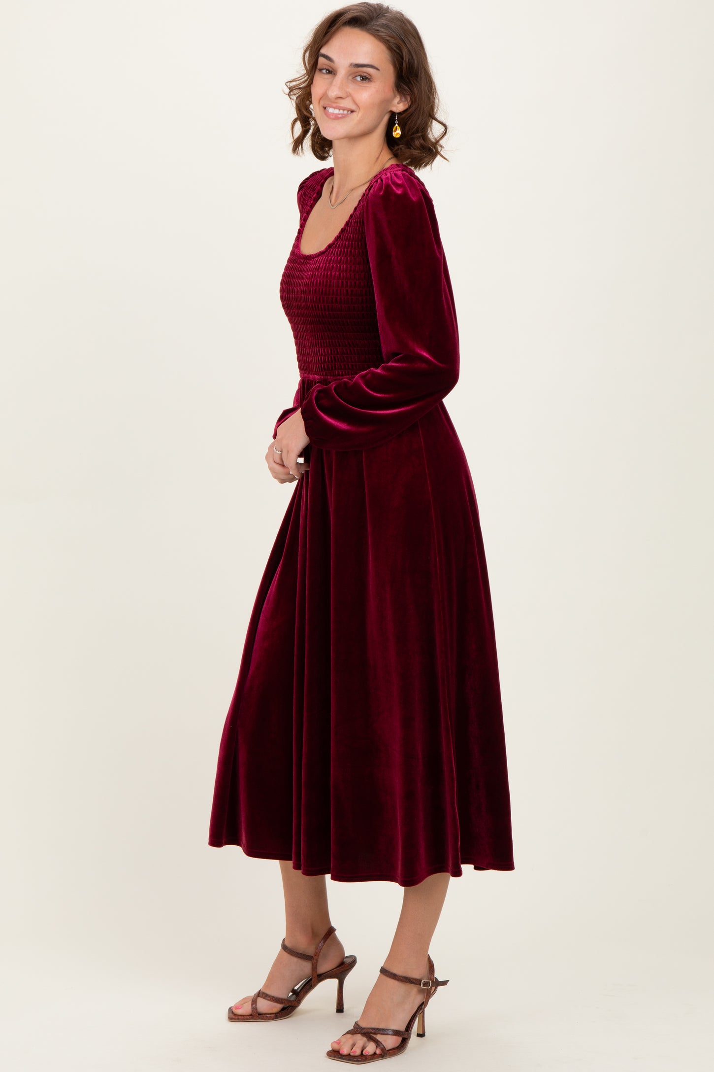woman wearing a burgundy velvet long sleeve dress with a smocked bodice and a flowing skirt, smiling and posing