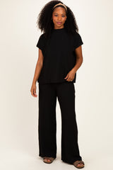 woman wearing a black ribbed short sleeve top and matching wide-leg pants, standing with one hand on her hip