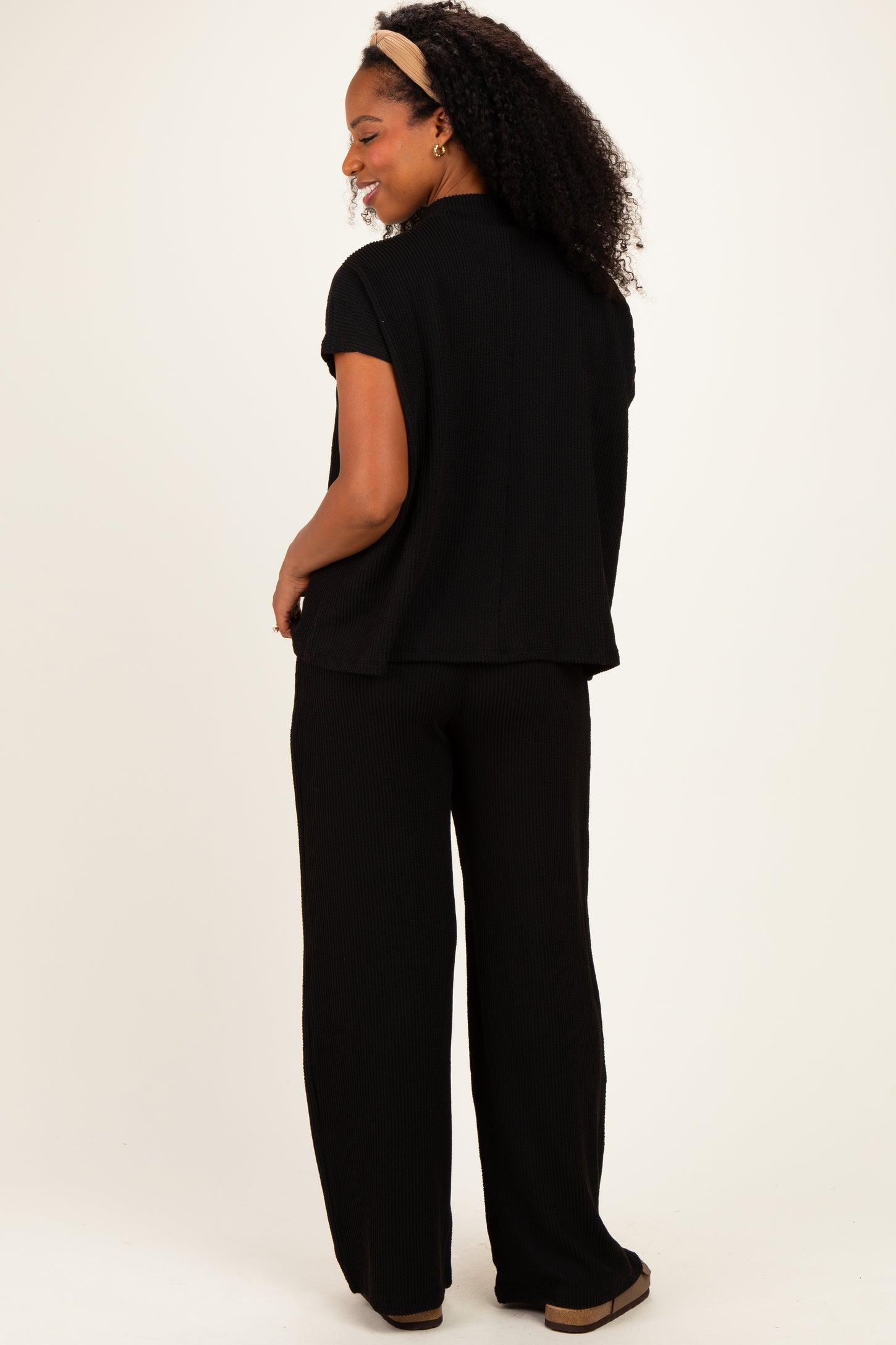 woman wearing a black short sleeve top and matching wide-leg pants, standing sideways with curly hair and a headband