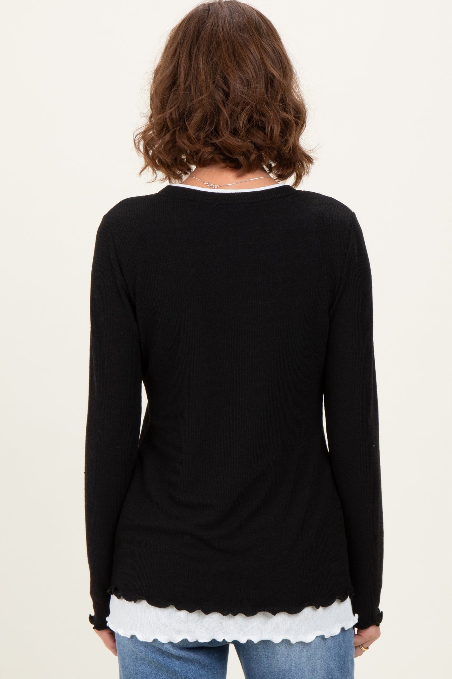 woman wearing a black long sleeve top with a scalloped hem layered over a white shirt, viewed from the back