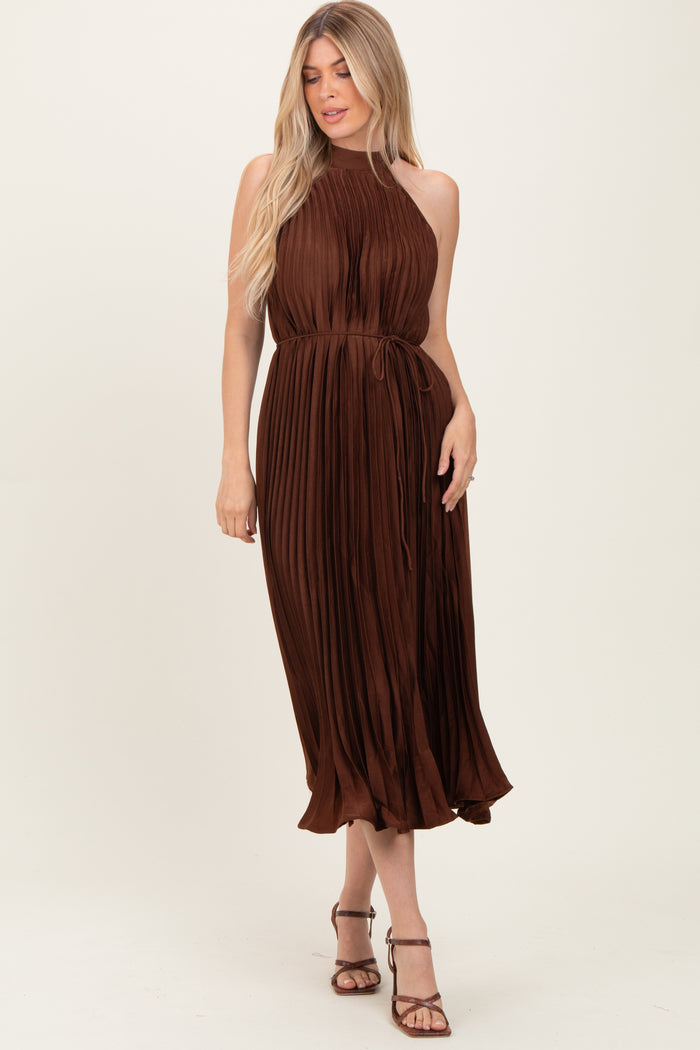 woman wearing a brown pleated midi dress with a halter neck and a tie waist, standing against a neutral background