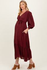 woman wearing a burgundy maternity dress with a tie waist, long sleeves, and a tiered skirt, displaying her baby bump