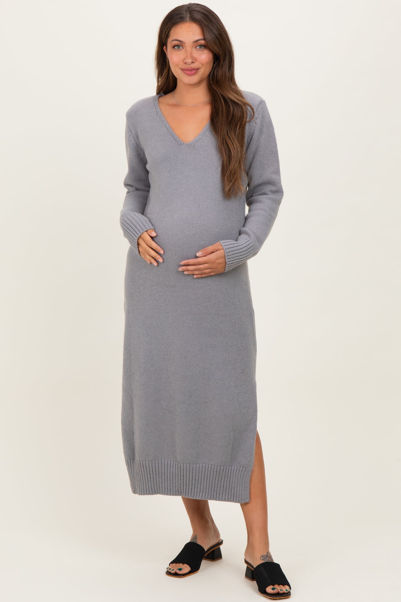 woman wearing a gray long sleeve maternity sweater dress with a v-neck and side slit, cradling her baby bump