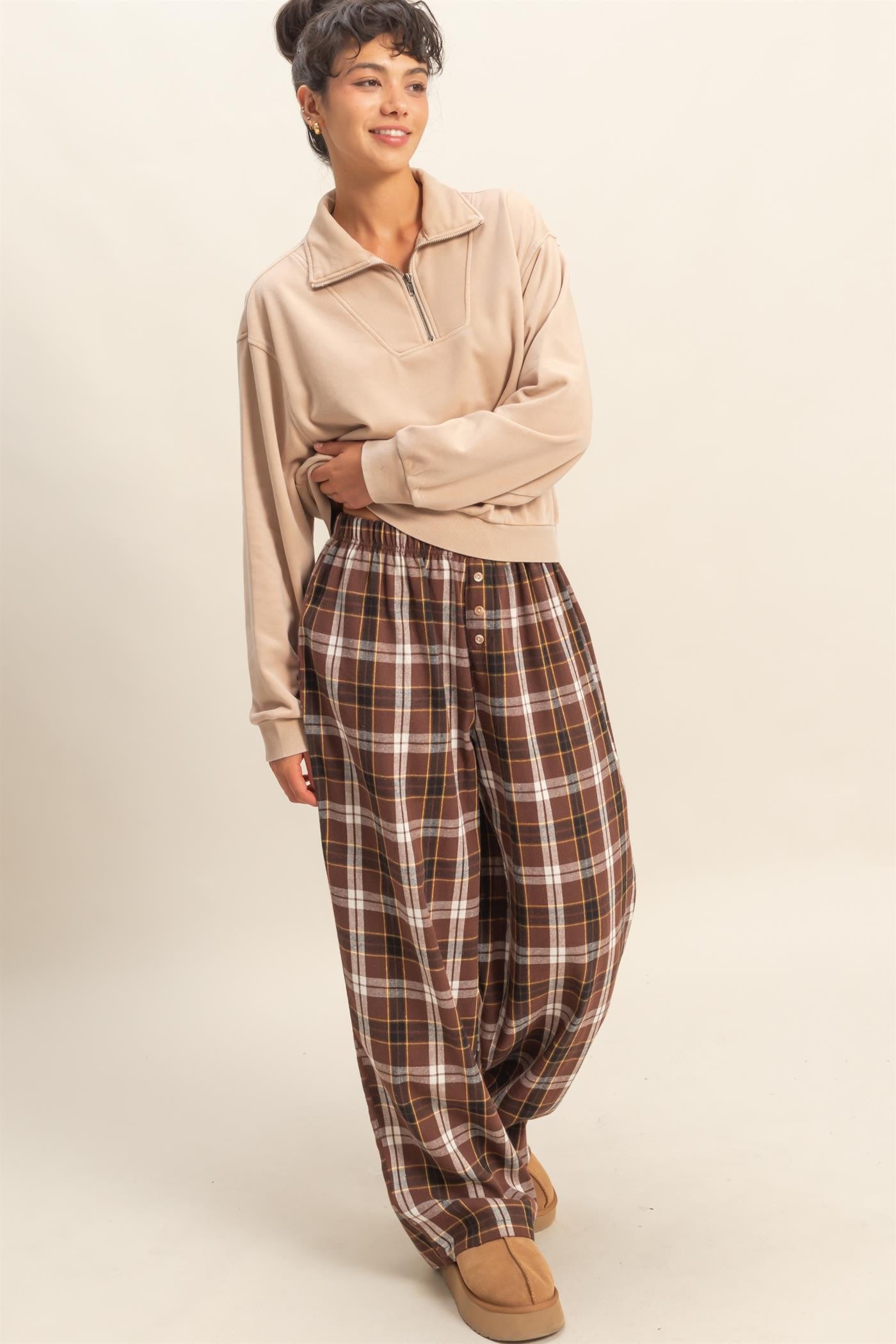 woman wearing a beige zip-up pullover and brown plaid wide-leg pants with buttons, posing in a relaxed manner
