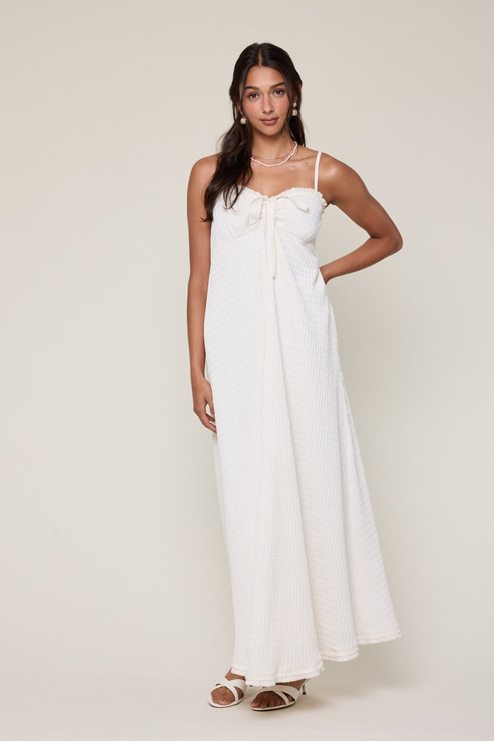 woman wearing a white maxi dress with bow detail and thin straps, standing casually with one hand on her hip