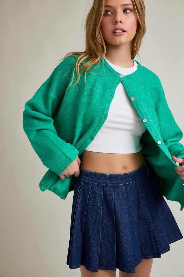 woman wearing a green cardigan over a white crop top and a denim pleated skirt