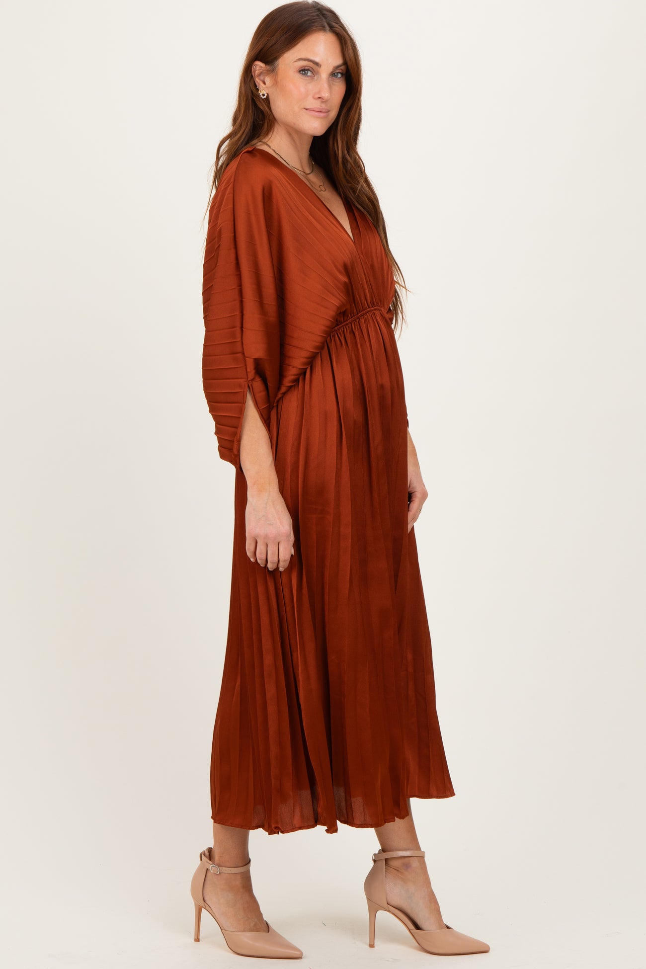 woman wearing a rust-colored pleated maxi dress with a deep v-neck and cinched waist, paired with beige heels