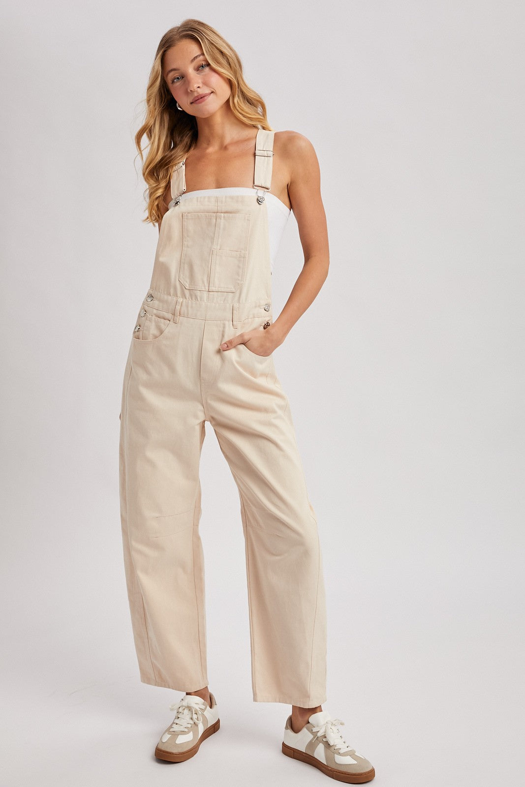 woman wearing a beige wide-leg overall with adjustable straps and front pockets, paired with a white top